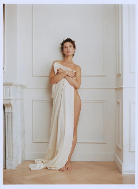 Portrait of a brunette adult woman draped in a cream-colored linen cloth in a white-walled room with hardwood floors and natural light.