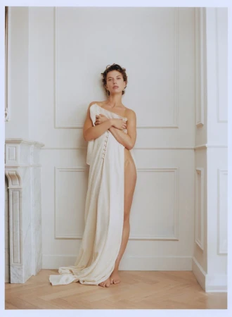 Portrait of a brunette adult woman draped in a cream-colored linen cloth in a white-walled room with hardwood floors and natural light.