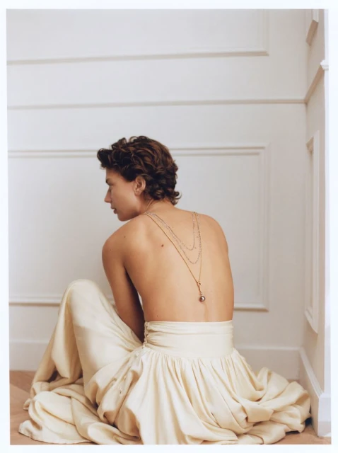Back view of a young adult woman with short brown hair sitting in a white dress with a gold chain necklace in a minimal interior.
