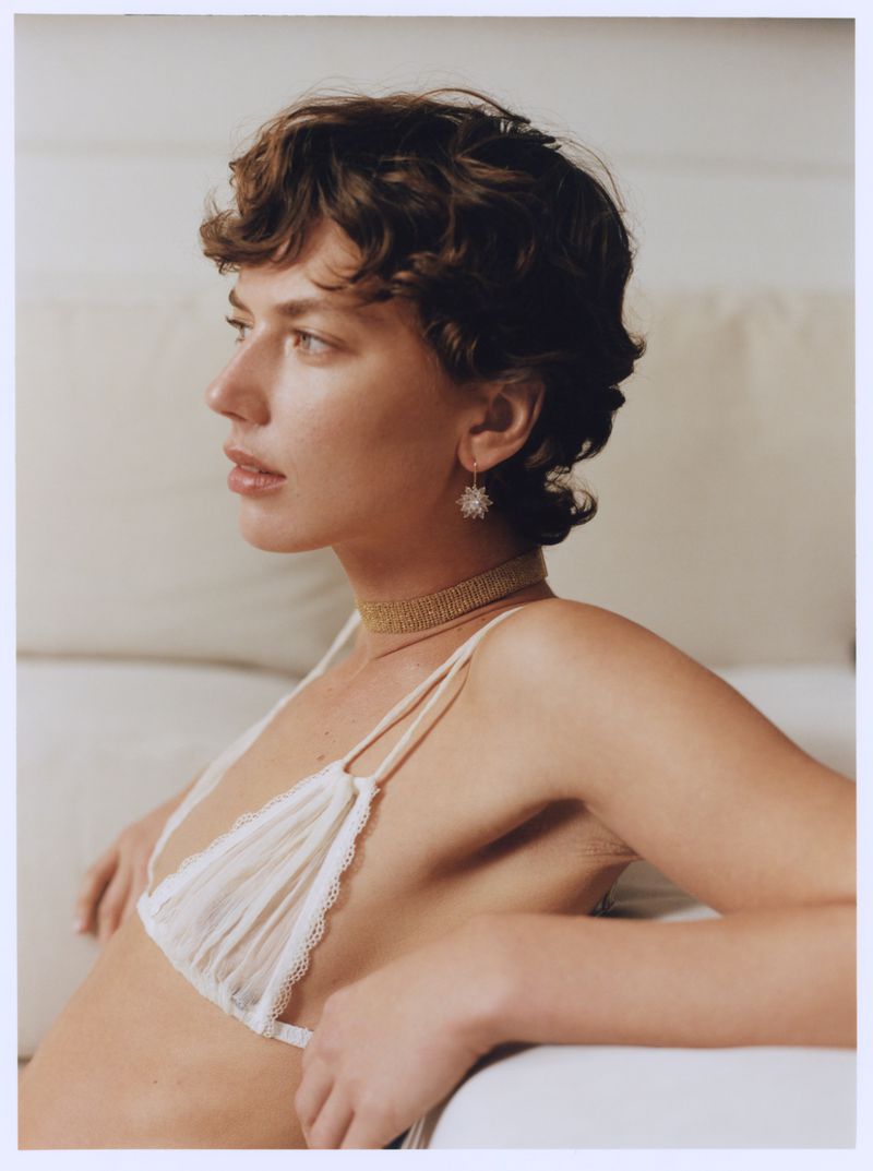 Portrait of a woman with short dark curly hair wearing a white lace bralette and delicate earrings against a soft neutral background