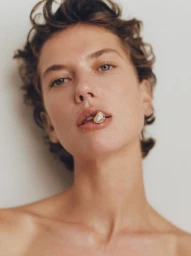 Close-up portrait of a model with short curly brown hair holding a gold ring in their mouth against a plain white background.