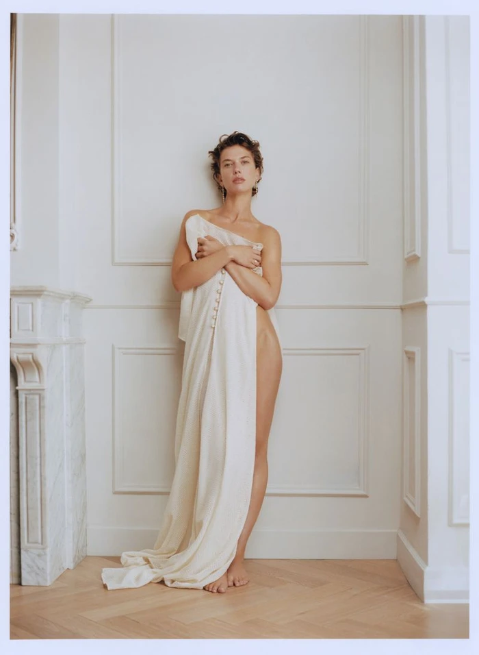 Model wrapped in a cream-colored fabric standing against a white paneled wall with a marble fireplace mantle to the side.