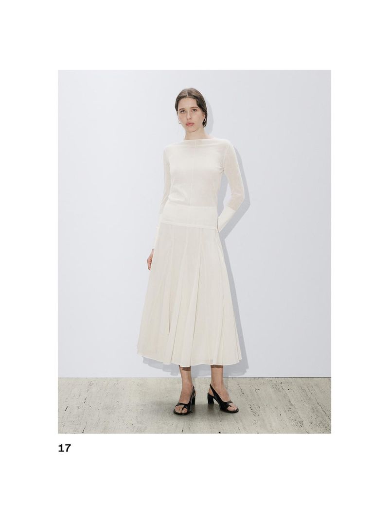 A slender woman poses indoors wearing a light cream-colored long-sleeve top with matching pleated skirt and black heeled sandals.