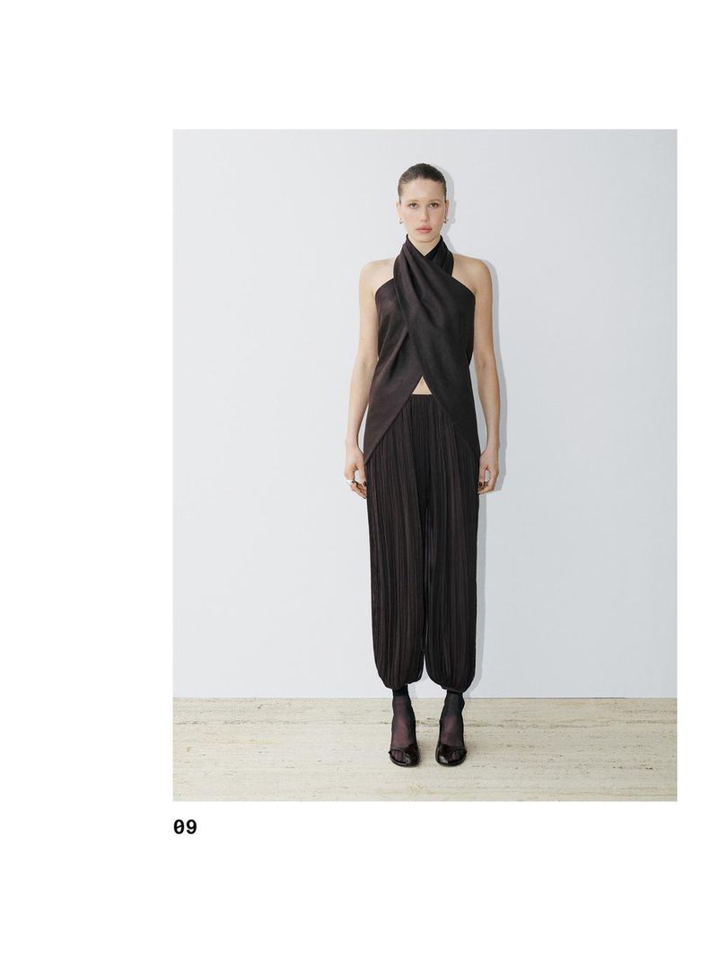 A slender fashion model poses in a dark brown, pleated top and matching pleated harem pants against a stark white backdrop.