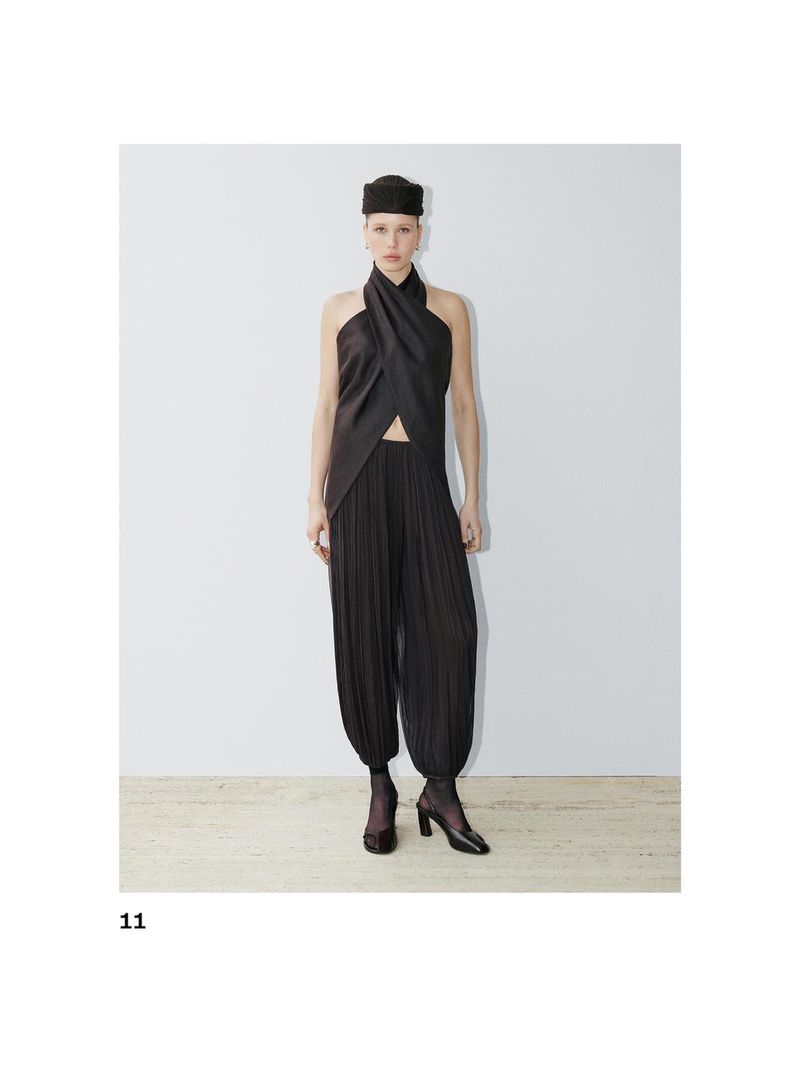 Fashion shoot of a woman in a dark halter top and pleated pants, posing against a white backdrop in a photo studio. Square format.