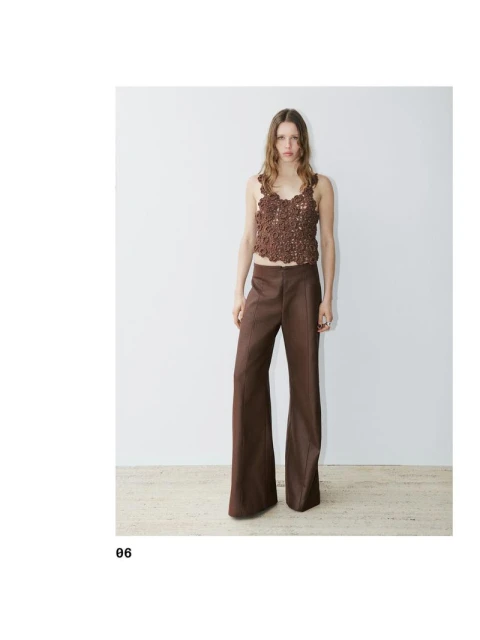 A slender woman is posing indoors wearing a brown, floral-patterned crochet tank top and brown, wide-leg pants against a white wall.