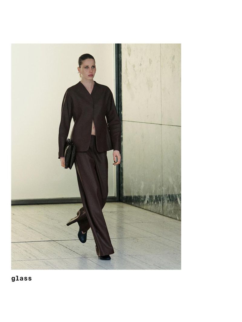 Model walking in a brown V-neck jacket and matching trousers holding a black clutch inside a modern hall