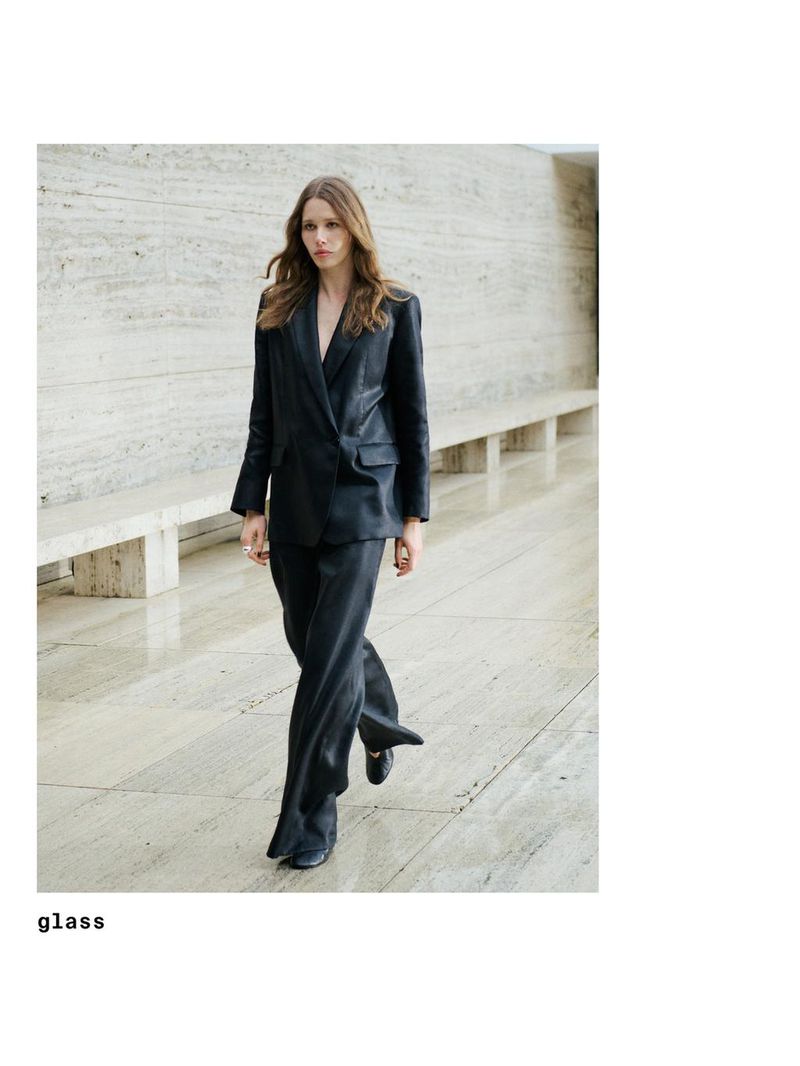 A woman wearing a black suit walks on a light grey tiled floor in a well-lit interior space with a minimalist design, showcasing fashion.