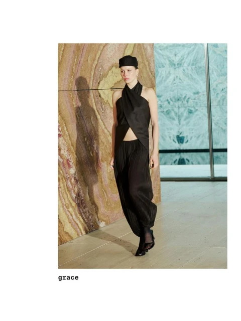 A woman in a black halter top and harem pants poses in front of marble walls, wearing a matching cap and heeled shoes.