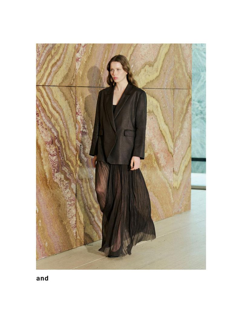 A woman in a dark blazer and sheer, pleated skirt poses against a textured wall in a fashion shot, showcasing elegant and modern design.