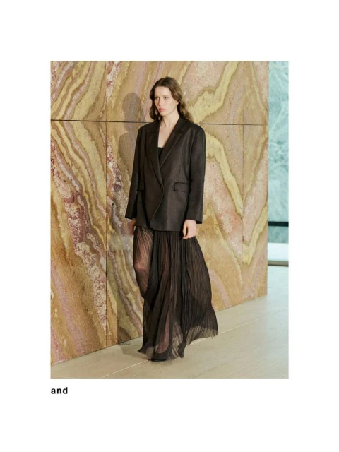 A woman in a dark blazer and sheer, pleated skirt poses against a textured wall in a fashion shot, showcasing elegant and modern design.