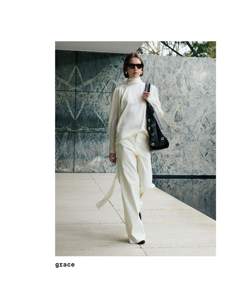 A woman in a white pantsuit and sunglasses walks outside with a black purse, showcasing a modern and fashionable style against a marbled background.