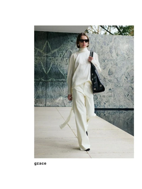 A woman in a white pantsuit and sunglasses walks outside with a black purse, showcasing a modern and fashionable style against a marbled background.