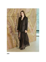 Model standing against a patterned marble wall wearing a dark oversized blazer and a long sheer pleated skirt
