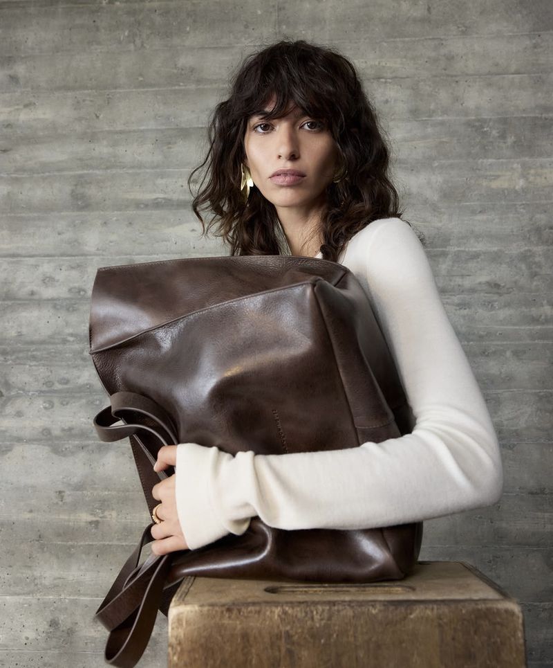A woman with dark curly hair, wearing a white sweater and gold earrings, stands with a brown leather backpack in a studio.