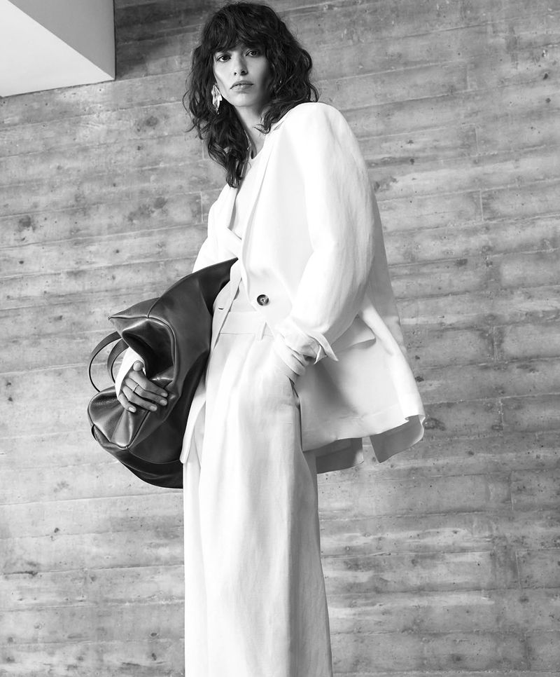 Model with dark wavy hair wearing white blazer and trousers, holding dark leather bag against textured concrete wall.