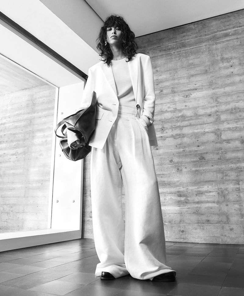 Monochrome fashion lookbook image of a model wearing a light-coloured blazer and wide-leg trousers while holding a large dark leather bag.