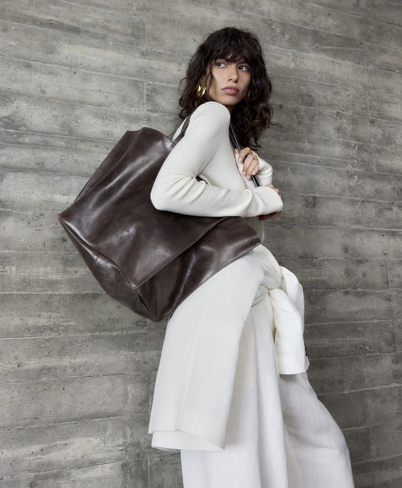 Woman in stylish white outfit poses with a dark brown leather tote bag against a textured gray wall, showcasing fashion and design.
