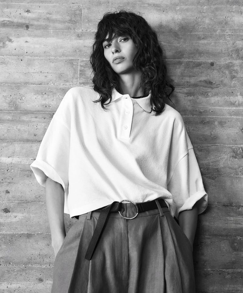Black and white portrait of a female model with curly hair wearing a white polo shirt, grey trousers, and a black belt against a grey wall.