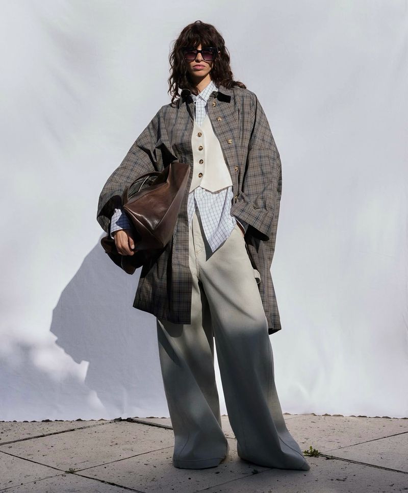 Model wearing a plaid coat, white vest, and wide-leg trousers holding a brown leather bag against a white seamless backdrop.