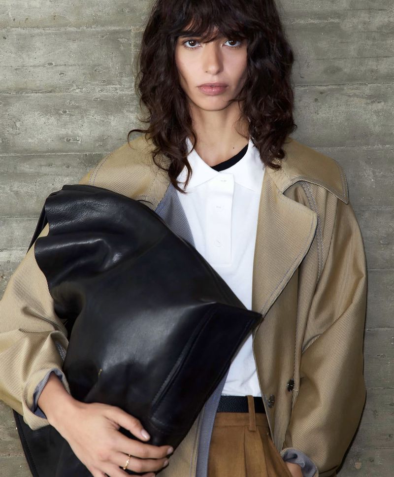 Model with dark wavy hair wearing a tan trench coat holding a large black leather bag against a concrete wall