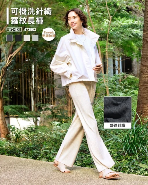 Fashionable woman in white jacket and linen pants walks on stone path surrounded by trees and greenery, showcasing relaxed style.