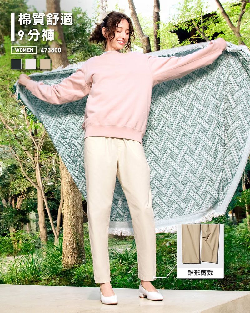 Woman in casual clothing holds patterned blanket, smiles outdoors surrounded by greenery. Light pink sweatshirt, beige pants, and white heels.