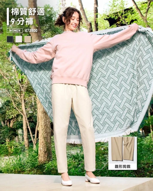 Woman in casual clothing holds patterned blanket, smiles outdoors surrounded by greenery. Light pink sweatshirt, beige pants, and white heels.