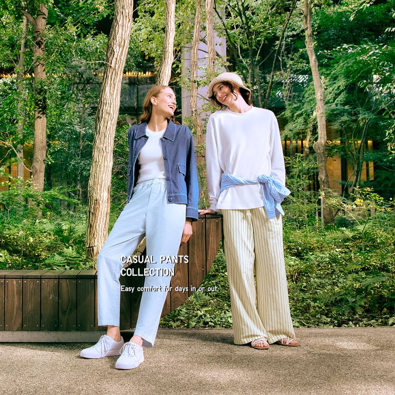 Two women pose in a casual fashion shoot outdoors in a wooded park, showcasing light-colored trousers and tops, surrounded by lush greenery and trees.
