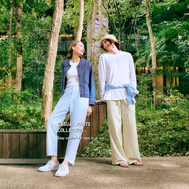 Two women pose in a casual fashion shoot outdoors in a wooded park, showcasing light-colored trousers and tops, surrounded by lush greenery and trees.