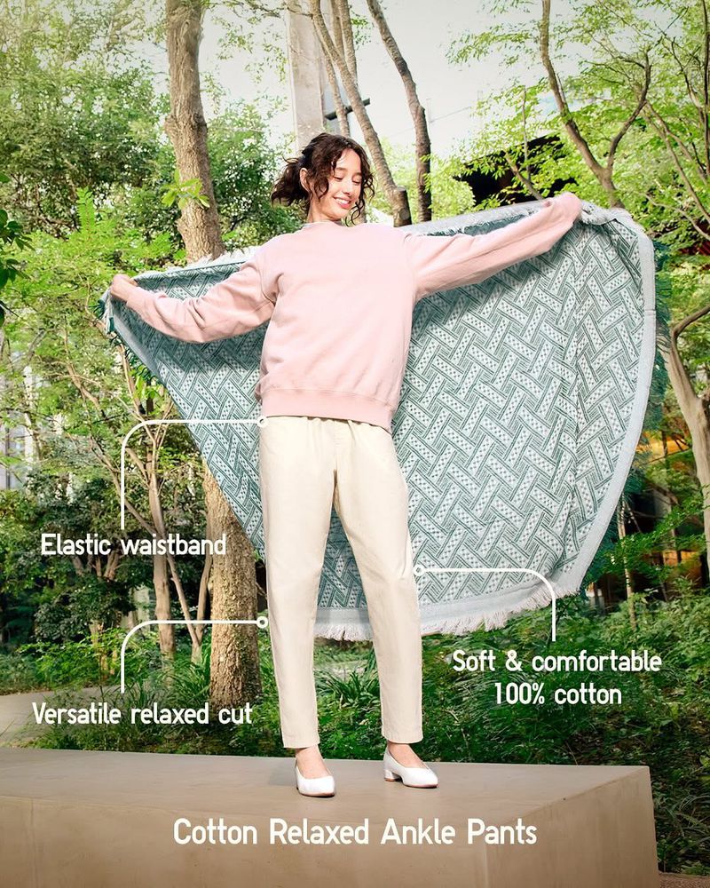 A young woman stands in a park-like setting holding a large, patterned blanket, wearing a pink sweatshirt, white pants, and white shoes.