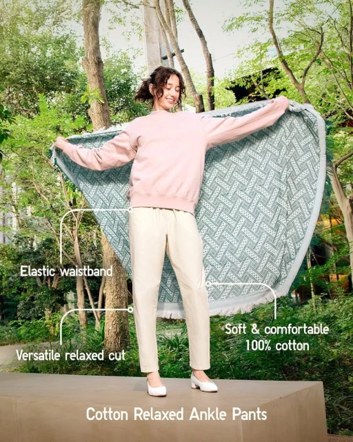 A young woman stands in a park-like setting holding a large, patterned blanket, wearing a pink sweatshirt, white pants, and white shoes.
