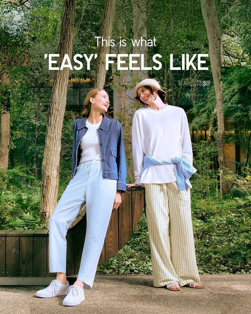 Two smiling women pose outdoors in a green, wooded setting, wearing casual summer clothes, light pants, and sneakers; style and fashion.