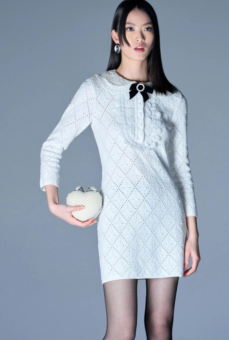 Young woman in a stylish white open-knit dress with a black bow, holding a beaded heart-shaped purse, studio fashion shot.