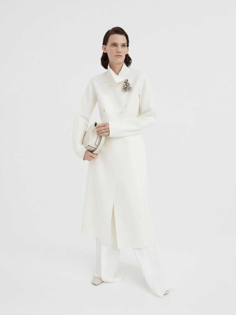 A woman in a white coat and pants stands in a white studio, holding a light-colored purse, posing for a full-length fashion portrait.