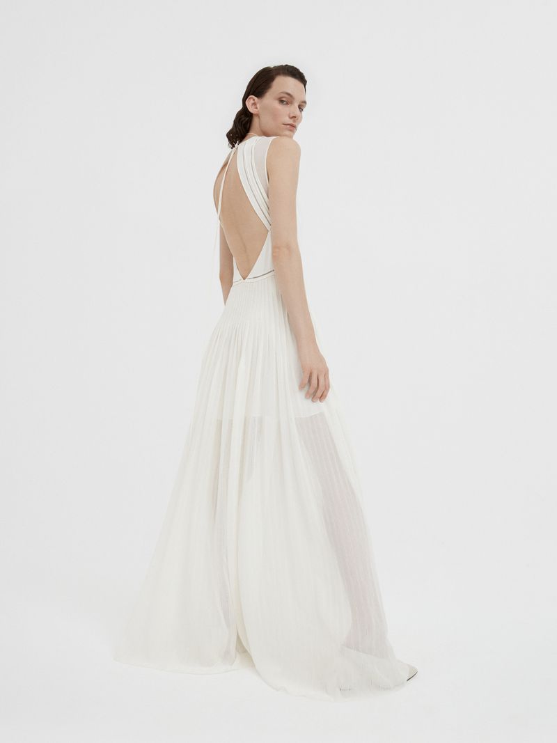 Model in a white sleeveless backless evening gown standing against a plain light studio background
