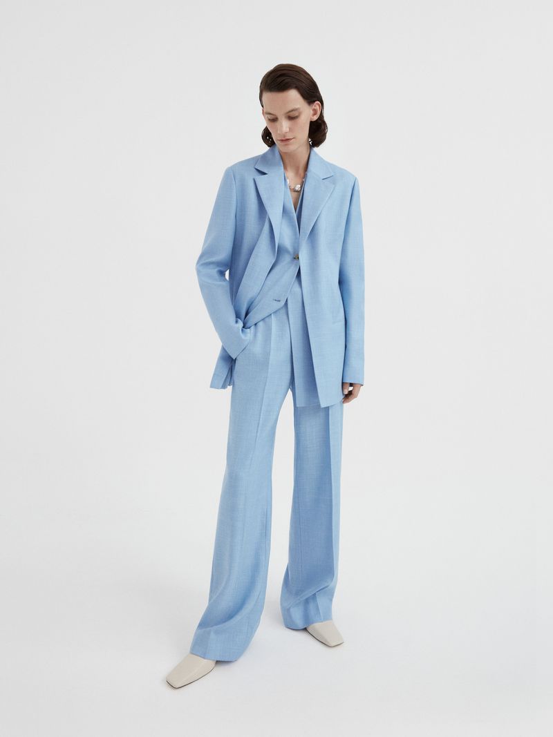 Woman in light blue blazer and wide-leg pants posing in a full shot against a white backdrop, minimalist fashion editorial photography.