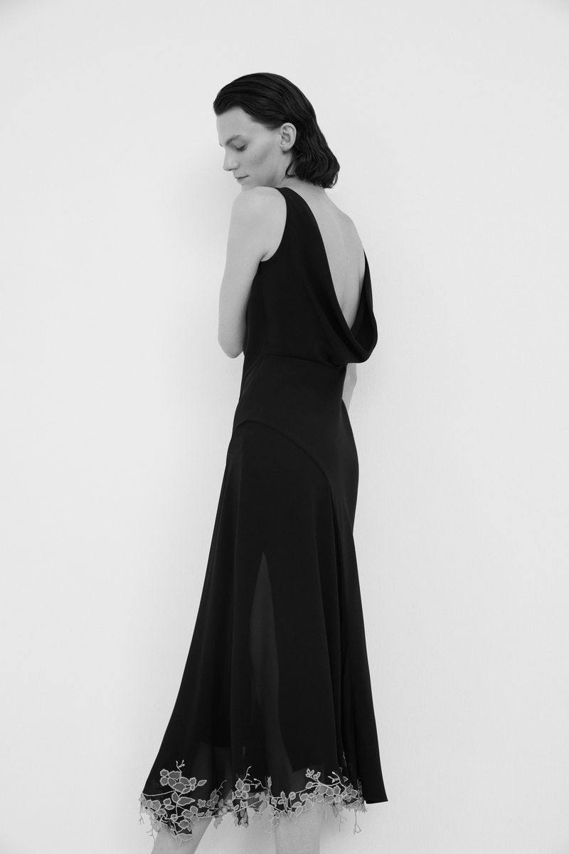 A woman with short dark hair models a low-backed black dress with a floral lace hem in a photo studio setting with soft, diffused lighting.