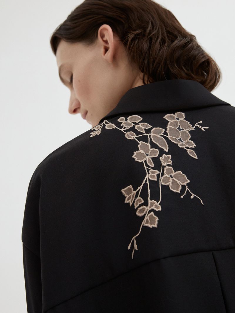 Woman with dark hair wearing a black jacket with embroidered floral design on the back. The background is a solid, neutral tone.
