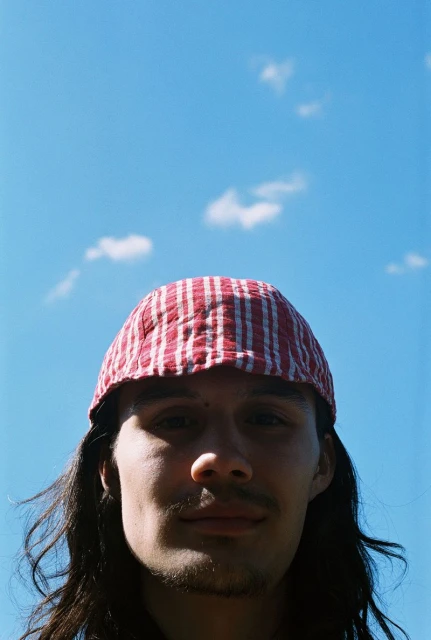 Eye-level close-up of a young adult with long dark hair wearing a red-and-white striped hat against a sunny blue sky with scattered clouds.