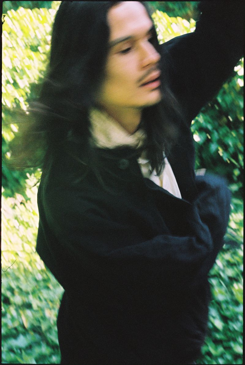 Portrait of a long-haired person wearing a black jacket and white shirt outdoors against a leafy green background.