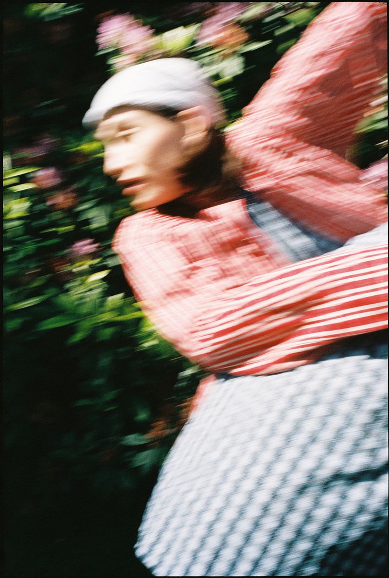 A blurry, slightly out-of-focus photo of an adult female wearing a hat and striped shirt, standing outdoors amid green foliage and pink flowers.