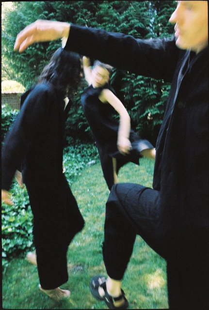 Three figures in dark clothing move across a lawn with greenery, captured in a dynamic, slightly blurred photograph emphasizing motion.