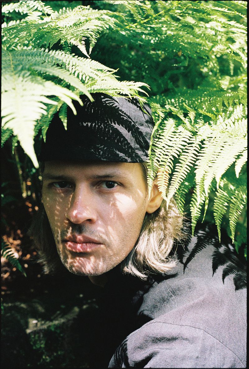 Close-up portrait of a man in a fern garden, wearing a patterned cap, with light shadows across his face and long blond hair.