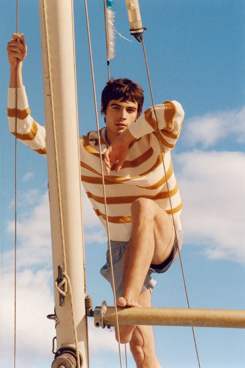 Young adult male poses on a sailboat wearing a striped top and shorts against a partly cloudy blue sky in a fashion editorial.