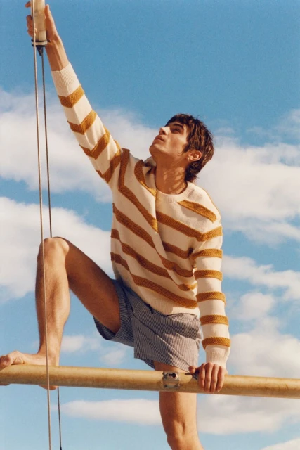 Man in a striped sweater and shorts reaches up on a sunny day, showcasing a casual fashion style with a bright, airy atmosphere.