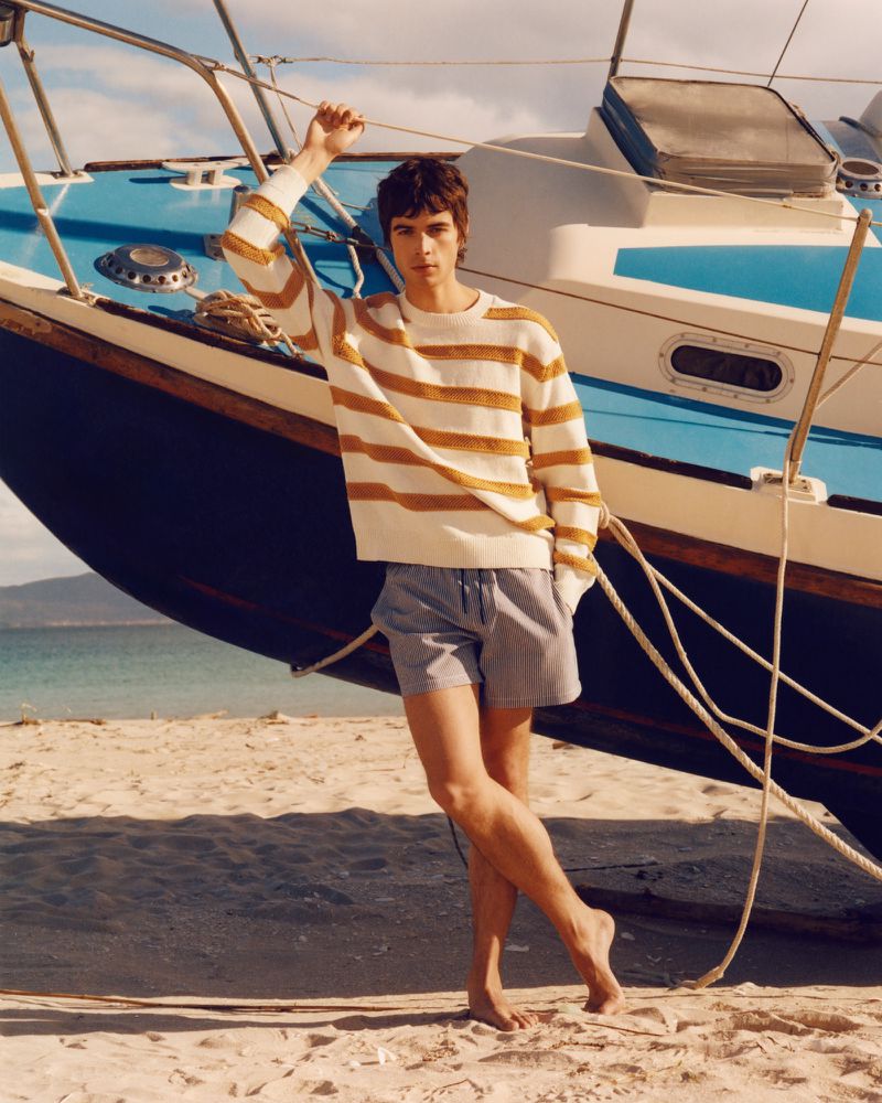 Young man standing barefoot on a sandy beach with a blue and white sailboat behind him, wearing a striped sweater and shorts.