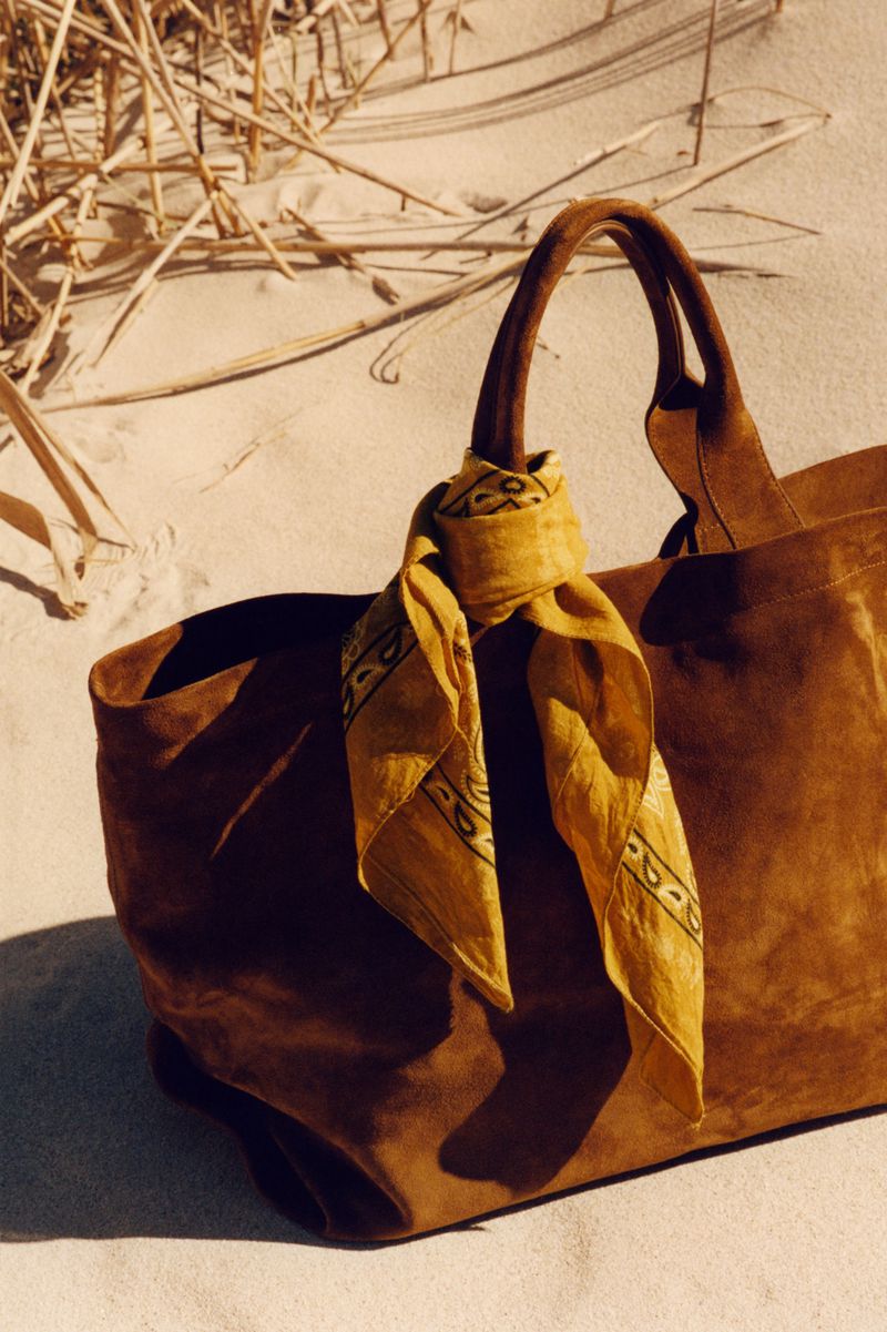 A large brown leather tote bag with a yellow patterned scarf tied to the handle rests on a sandy beach under bright sunlight.