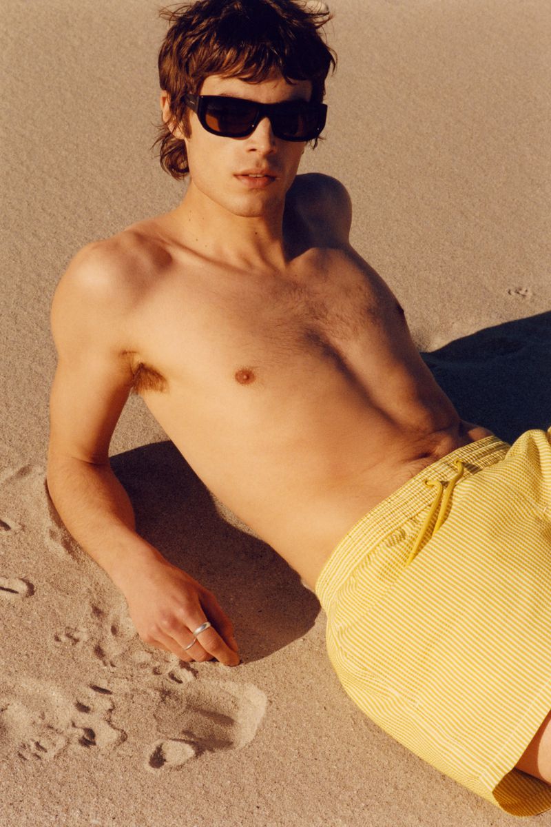 Young adult male model poses on a sandy beach wearing sunglasses and yellow swim trunks in an outdoor fashion lifestyle shot.