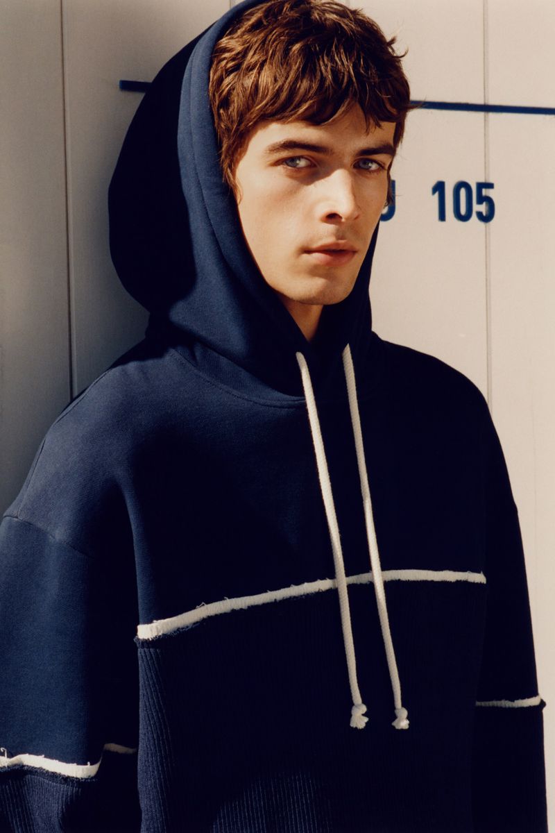 Close-up of a young adult male wearing a dark blue hooded sweatshirt, posed against a light-colored wall, looking directly at the camera.